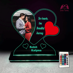 A custom acrylic LED lamp photo frame with heart-shaped panels, illuminated with multicolored lights, displaying a couple's photo. A remote control with various colors is also shown.