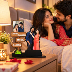 Personalized wooden sign couple memorial plaque displayed on a bedside table in a bedroom setting, with a couple in bed blurred in the background. The sign features custom couple photos, names 'ANURAG' and 'RADHIKA', and directional signs reading 'Endless Connection'.